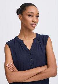 Blouse sans manches bleu marine avec un col, fermeture à boutons sur le devant et texture froissée. Fabriquée dans un tissu doux et fluide.