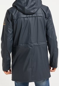 Veste imperméable bleu marine avec capuche, présentant une texture lisse, une taille ajustable et un logo doré sur l'épaule gauche.