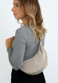 Beige axelremsväska med rundad form, gjord av mjukt tyg, med en slät yta och subtil branding. Bärs över kroppen med en grå topp.