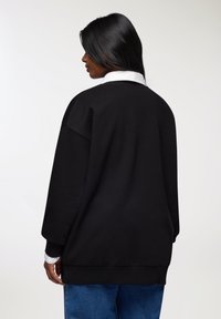 Felpa nera oversize con collo rotondo e spalle scese, abbinata a jeans blu. Texture liscia, design minimale.