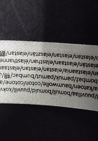 White textile label with black printed text, featuring repetitive pattern and a textured surface, attached to a black background.