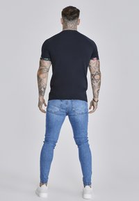 Marineblauw shirt met een aansluitende pasvorm en bloemenaccenten op de mouwen, gecombineerd met lichtblauwe skinny jeans en witte sneakers, gezien van de achterkant.