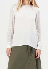Blouse blanche à manches longues transparentes texturées avec un motif en chevron, associée à une jupe drapée de couleur vert olive.