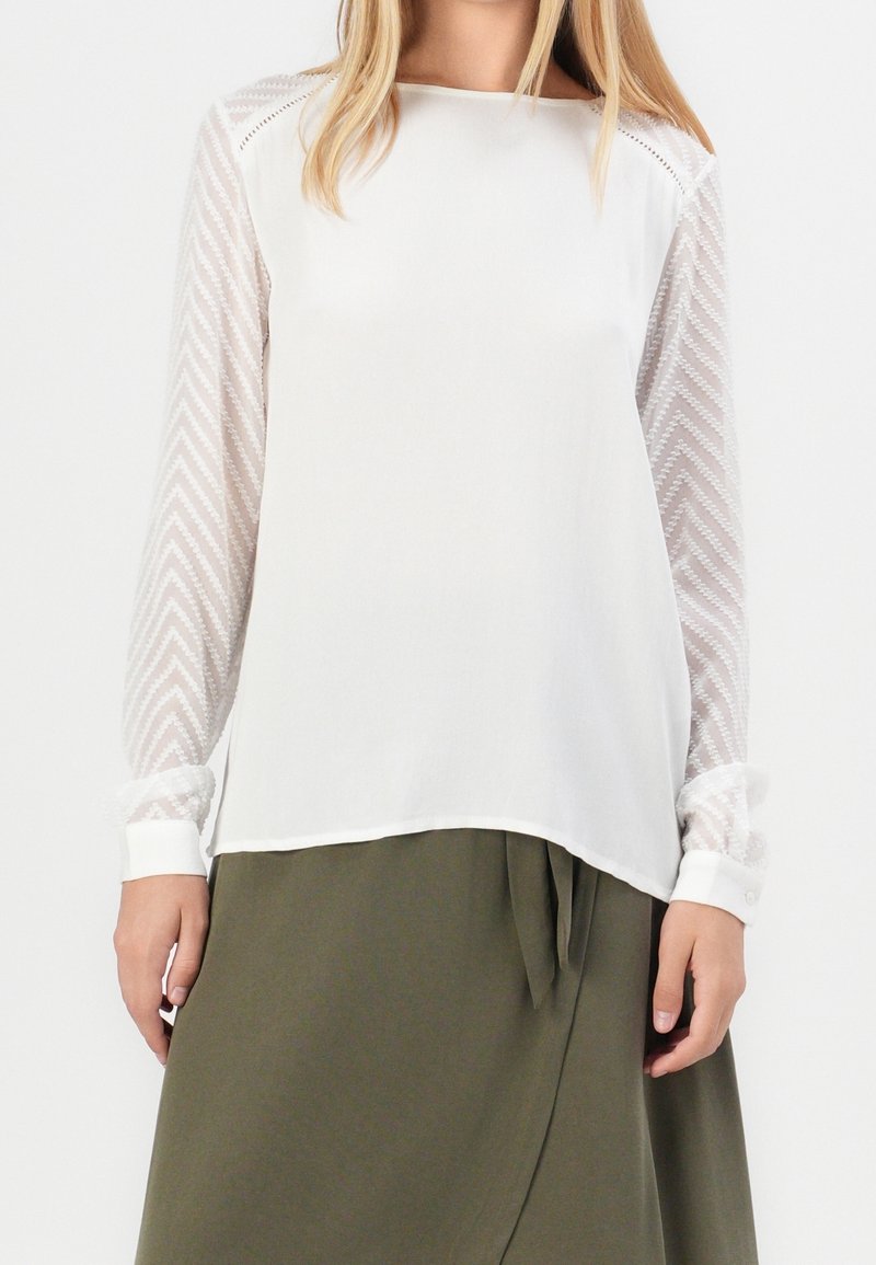 Blouse blanche à manches longues transparentes texturées avec un motif en chevron, associée à une jupe drapée de couleur vert olive.