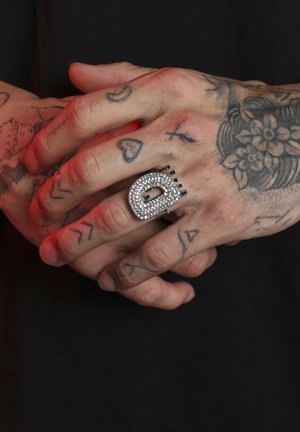 Anillo de plata incrustado de strass en forma ovalada y audaz, con acentos negros y detalles texturizados prominentes, que se lleva en una mano tatuada.