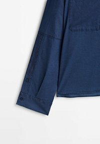 Chemise bleu marine à manches longues avec poignets boutonnés et détails de couture visibles, présentée sur un fond blanc uni.