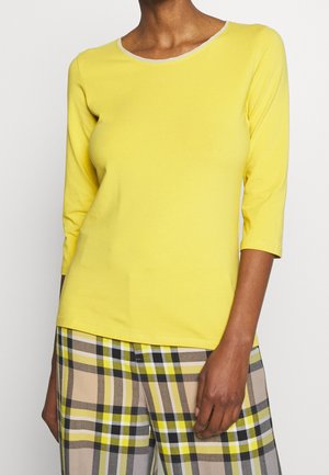 Persoon die een geel shirt met lange mouwen en ronde hals draagt en een geel, zwart en beige geruite broek, staand tegen een effen achtergrond.
