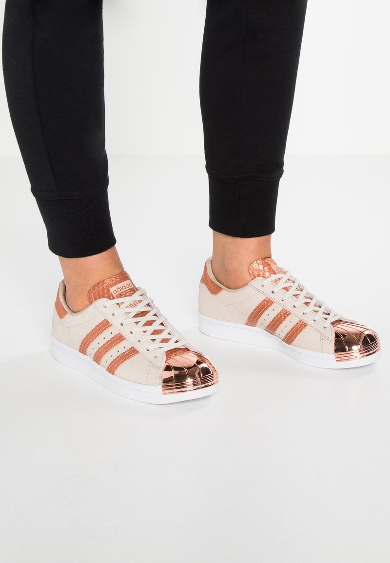 Sneakers with beige suede upper, rose gold accents, three stripes, and metallic shell toe. White rubber sole and flat laces.