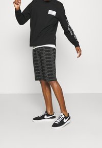 Czarna koszula z długim rękawem z białym logo Calvin Klein, w połączeniu z wzorzystymi czarnymi szortami z powtarzającą się nazwą marki i czarnymi sneakersami.