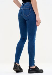 Vaqueros ajustados de mezclilla azul oscura, de talle alto y textura suave. Presentan bolsillos traseros y se combinan con zapatos negros de cordones.