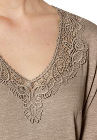 Beige Oberteil mit tiefem V-Ausschnitt und aufwendiger, lace-ähnlicher Stickerei in floralem Muster entlang des Ausschnitts. Weicher, strukturierter Stoff.