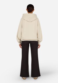 Világos bézs kapucnis pulcsi, lelógó vállakkal és bordás mandzsettával, fekete harangszárú nadrággal párosítva. Sima szövet textúra és laza szabás.