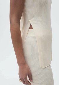 Ribbet strik top og nederdel sæt i lys beige, med sideslids, blød tekstur og tætsiddende design. Hånd viser et guldringe tilbehør.