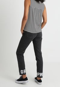 Czarne dżinsy o prostym kroju z wyraźnym białym napisem "GR" i "PW" na mankietach, w zestawie z pasiastym topem na ramiączkach i czarnymi trampkami.