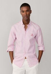 Homme portant une chemise à rayures roses et blanches à boutons, manches retroussées, debout les mains dans les poches devant un fond uni.