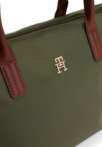 Grüne Nylon-Umhängetasche mit braunen Ledergriffen, versehen mit einem goldenen Logo und einem Reißverschluss. Glatte Textur, strukturiertes Design und minimalistische Details.