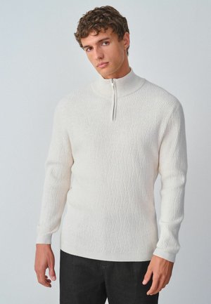 Hvid strikket sweater med høj krave, der har en diamantmønstertekstur og en halvlynlås ved halsen.
