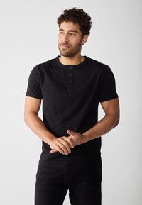Svart kortermet henley-skjorte laget av bomull med rund hals, med tre knapper og en tettsittende design. Båret med mørke jeans.
