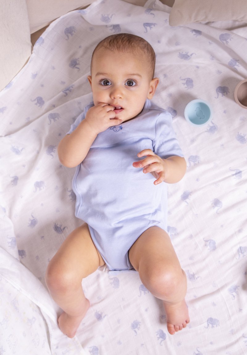 Hellblaues kurzärmliges Bodysuit an einem Baby, das auf einer gemusterten weißen Decke mit hellblauen Elefantendesigns liegt, umgeben von zwei kleinen Schalen.