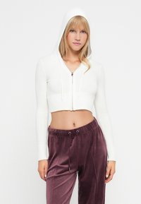 Junge Frau in einem weißen, kurzen Hoodie mit hochgestellter Kapuze und lila samtigen Jogginghosen vor einem schlichten weißen Hintergrund.