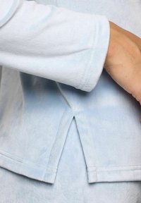 Prim-plan al unui mânecă de îmbrăcăminte din fleece de culoare bleu deschis, cu detalii de fanta laterală și cusături vizibile, iar o mână este parțial vizibilă în partea dreaptă.