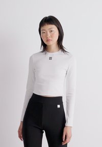 White long-sleeve cropped top with a crew neck, featuring a small black "HUGO" logo on the chest, paired with black high-waisted leggings.