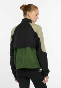 Vrouw met haar in een knot, gekleed in een lichtgewicht jack in kleurblokken zwart, olijf en groen, en zwarte leggings, die met haar rug naar de camera staat tegen een witte achtergrond.