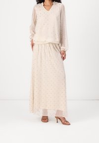 Ensemble deux pièces couleur crème composé d'un chemisier à manches longues et col en V, et d'une jupe maxi assortie ornée de motifs ponctués métalliques éparpillés.