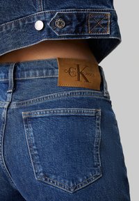 Calvin Klein džinsai su odiniu logotipu, tamsiai mėlynos džinsinės medžiagos, matomomis siūlėmis ir užpakalinėmis kišenėmis. Savybėms priklauso metalinės detalės ir tekstūruota medžiaga.