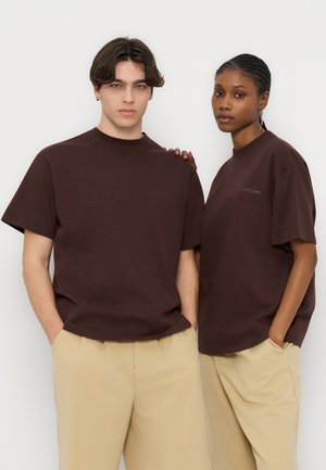 Giovane uomo e donna che indossano t-shirt marrone scuro abbinate e pantaloni beige, in piedi vicini con le mani nelle tasche contro uno sfondo bianco.
