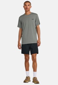 Camiseta atlética gris de manga corta con logo, combinada con pantalones cortos negros y zapatillas beige. Modelo de pie, con las manos relajadas a los lados.