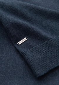 Tmavě námořnický pletený svetr s ribbovaným manžetem a rytým stříbrným štítkem s textem "ombre". Hladká textura s jemným leskem.