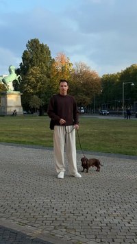 Hnedý sveter, krémové voľné nohavice a biele tenisky. Vedľa stojí malý hnedý pes na vôdzke. Kamenná dlažba a zelené stromy.
