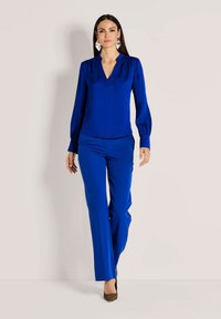 Blaue Seidenbluse mit V-Ausschnitt und langen Ärmeln, kombiniert mit passenden blauen maßgeschneiderten Hosen. Leopardenmuster-Absätze runden das Outfit ab.