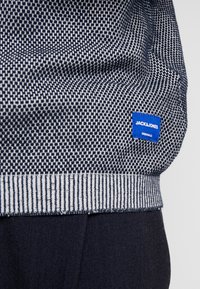 Marineblaues und weißes gemustertes Textil mit geripptem Saum, das ein blaues Stoffetikett mit der Aufschrift "JACK & JONES" und "ORIGINALS" zeigt.