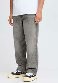 Hombre vestido con jeans sueltos de color gris desgastado, camiseta blanca, pulsera plateada y zapatillas bajas negras con cordones blancos y suelas color crema.