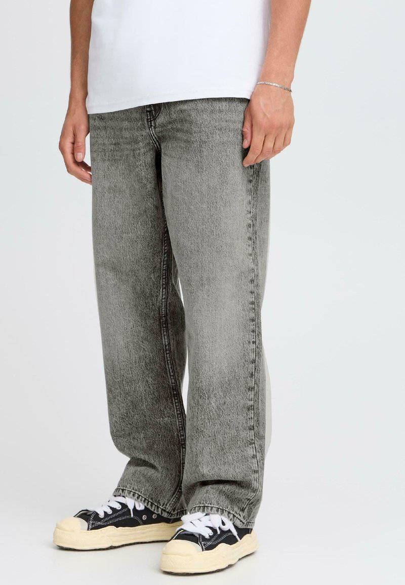 Hombre vestido con jeans sueltos de color gris desgastado, camiseta blanca, pulsera plateada y zapatillas bajas negras con cordones blancos y suelas color crema.