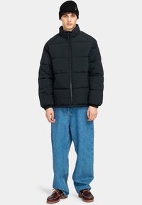 Giacca nera imbottita con colletto alto, chiusura con zip e sezioni cucite, indossata sopra jeans blu larghi e scarpe marroni con lacci. Cappello beanie nero.
