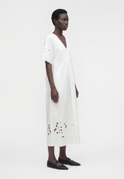 Femme debout dans une robe midi blanche avec des détails œillets sur les manches et l'ourlet, portant des chaussures plates noires à bout pointu, sur un fond uni.