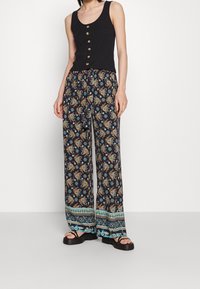 Černý žebrovaný top na ramínka se zlatými knoflíky, zkombinovaný s námořnickými širokými kalhotami se vzorem s květinovými motivy a modrými akcenty na lemu.