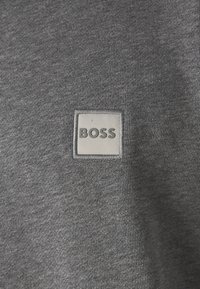 Sötét szürke pulóver puha anyagból, elöl egy négyzet alakú fehér címkével, amelyen vastag szürke betűkkel a "BOSS" felirat látható.