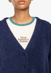 Marineblauwe fuzzy cardigan met V-hals, voorzien van zwarte knopen. G gedragen over een lichtcrème shirt met turquoise rand en geborduurd logo.