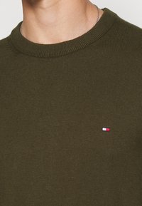 Oliwkowy dzianinowy sweter z prążkowanym, okrągłym dekoltem i małym haftowanym logo z akcentami w kolorach czerwonym, białym i niebieskim na lewej piersi.