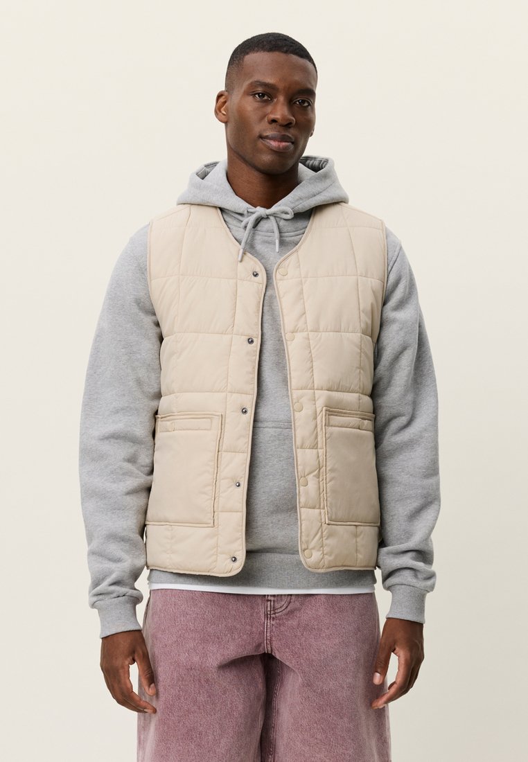 Μπεζ γιλέκο με καπιτονέ σχέδιο, κουμπωτό με κουμπιά αστραπή και δύο εμπρόσθιες τσέπες, φορεμένο πάνω από μια γκρι φούτερ με κουκούλα. Το μοντέλο έχει ουδέτερη έκφραση και στέκεται μπροστά σε ανοιχτό φόντο.