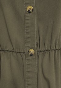 Olivgrön tyg med en mjuk textur, utrustad med två glänsande gyllene knappar och veckad elastik i midjan.