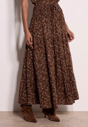 Femme portant une jupe longue marron à fleurs avec une taille smockée, associée à des bottines marron en daim à bout pointu.