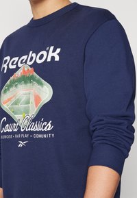 Marinblå sweatshirt med ribbade muddar och hals. Har en grafik av en tennisbana med berg och stadslandskap. Texten inkluderar "Reebok Court Classics."