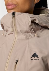 Lys beige jakke med høy krage, med glidelås foran og snorer. Viser en svart fjelllogo på brystet.