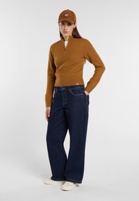 Brown zip-neck sweater, dark blue wide-leg jeans, and white shoes. The cap matches the sweater in color with a logo detail on the front.