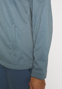 Gray, textured pullover with a zip collar and side pocket. Features a fitted sleeve and seamless shoulder design. Material is soft and breathable.